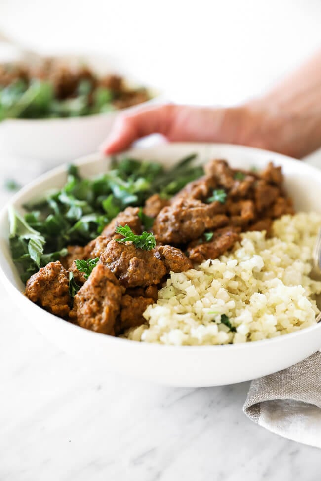 Angled image of setting a bowl of easy curry beef with cauliflower rice and greens down on the table. 