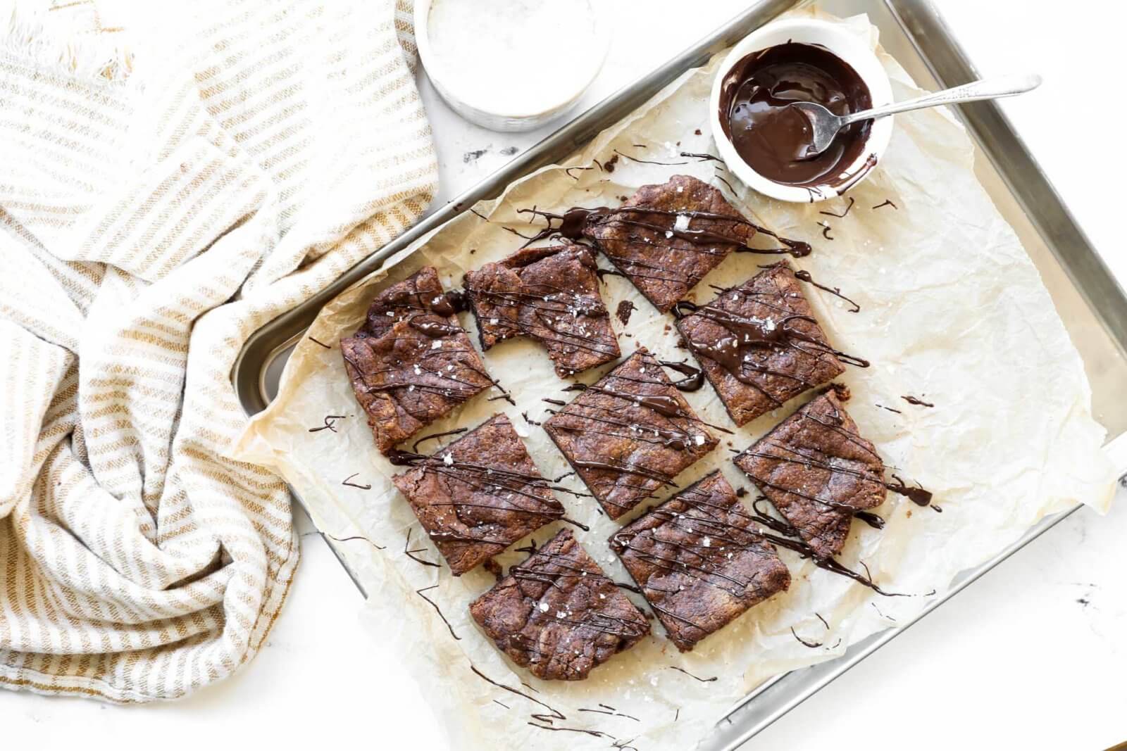 Horizontal overhead image of dairy free brownies sliced into squares on a sheet pan. Drizzled with melted chocolate and sprinkled with flaky sea salt. 