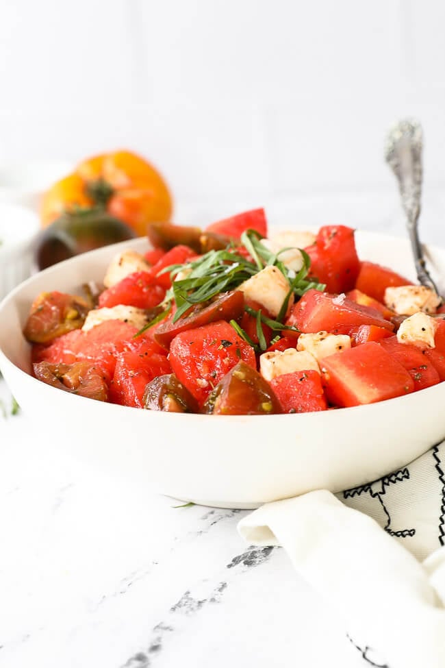 Straight on image of tomato watermelon salad in a bowl with a serving spoon. Fresh basil on top. 