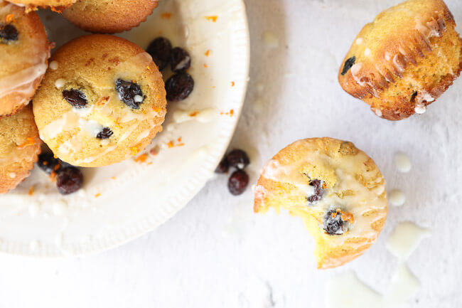 Horizontal image of gluten free orange cranberry muffins. Some on a plate. One on the table next to plate with a bite taken out of it. Orange icing drizzled on top. 