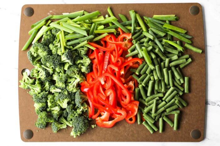 Prepped broccoli florets, green beans, red bell pepper and sliced onions on a cutting board