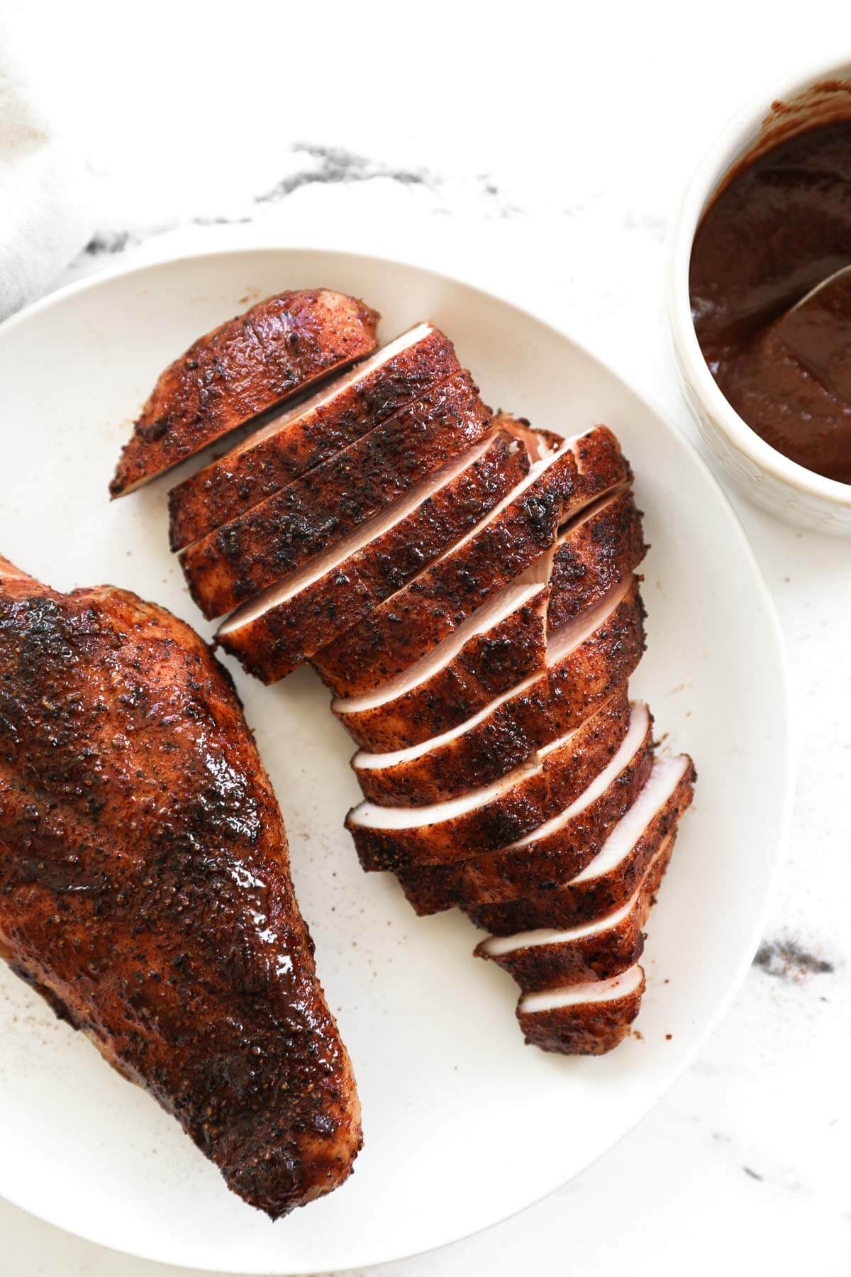 Two pieces of smoked chicken breast on a plate. One piece sliced into strips. BBQ sauce in a small bowl on the side.
