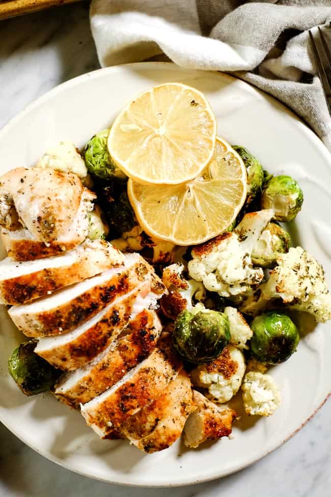 Closer up shot of easy lemon chicken served on a plate with roasted brussels and cauliflower and garnished with lemon slices. 