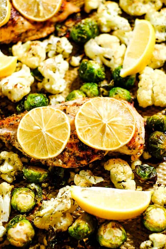Close up angled shot of easy lemon chicken and roasted brussels and cauliflower on sheet pan with lemon slices. 