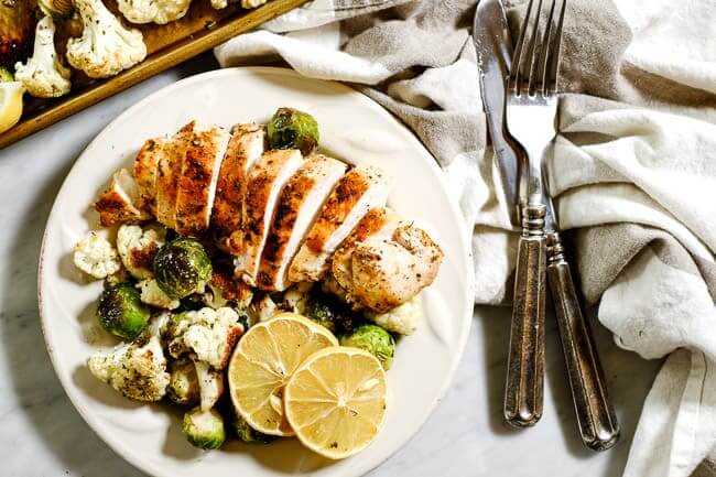 Easy lemon chicken served on a plate with roasted brussels and cauliflower and lemon slices for garnish, a grey and white striped napkin and silverware. 