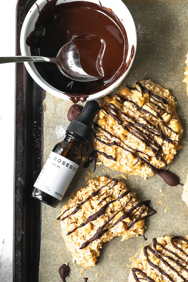 No bake cbd cookies on a cookie sheet drizzled with chocolate on top. One cookie with a bite taken out of it. Bottle of Rosebud CBD oil on the cookie sheet. 