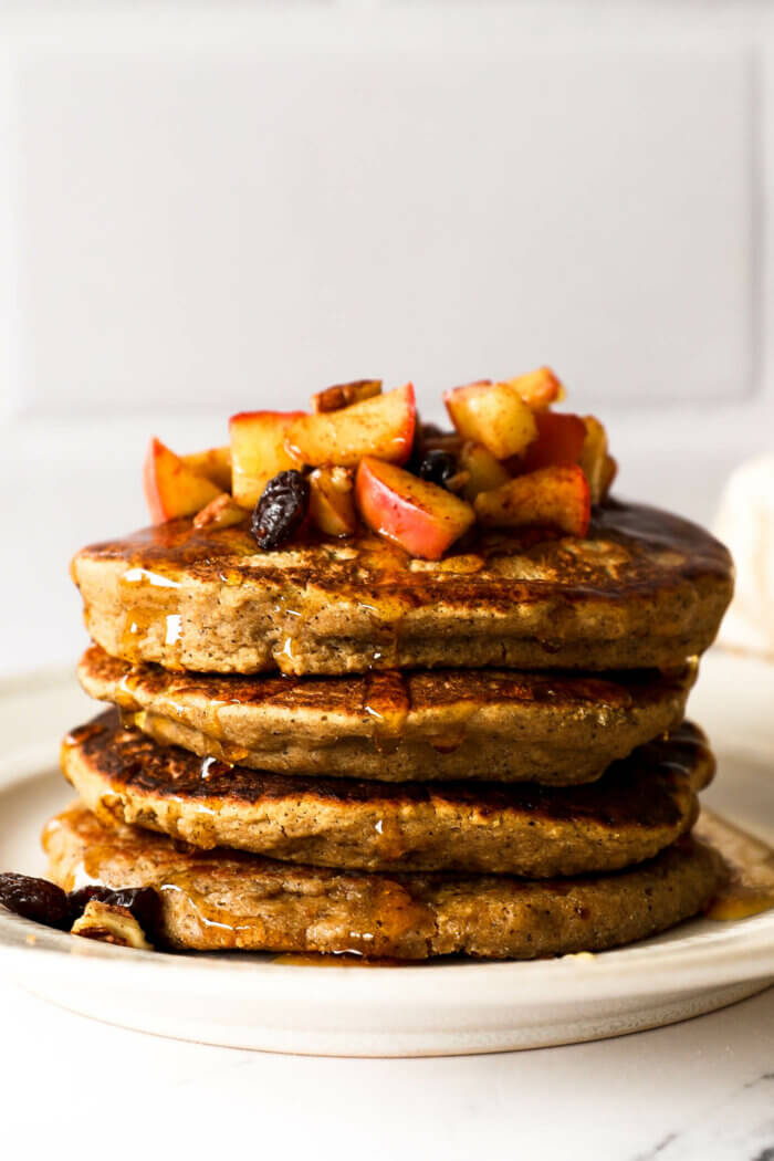 A stack of pancakes on a plate. Topped with sauteed apples, raisins and pecans with a pour of maple syrup over the top.
