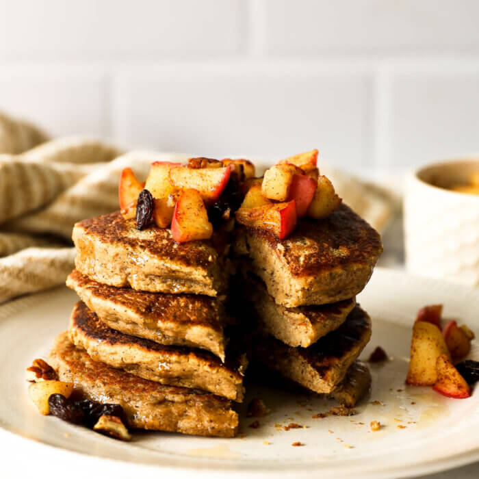 A stack of pancakes on a plate with a slice taken out. Topped with sauteed apples, raisins and pecans with a pour of maple syrup over the top.