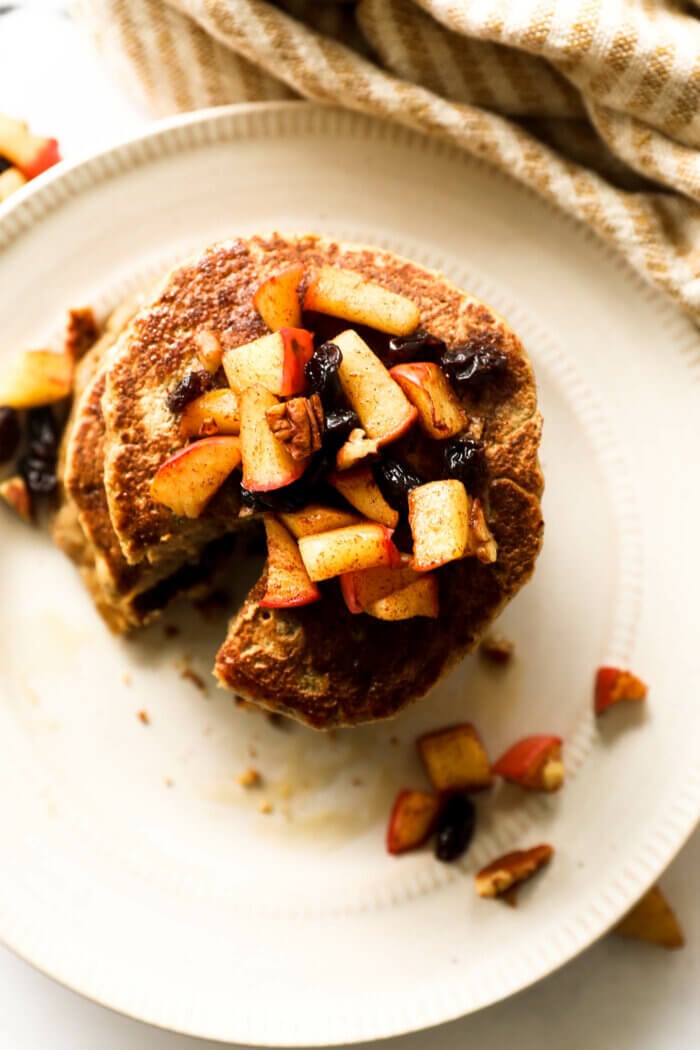 Overhead image of a stack of pancakes with an apple, raisin and pecan topping. All with maple syrup poured over the top.