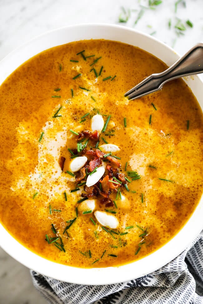 Vertical overhead close up image of pumpkin soup in a bowl with a spoon. Topped with bacon bits, pumpkin seeds and chopped chives. 