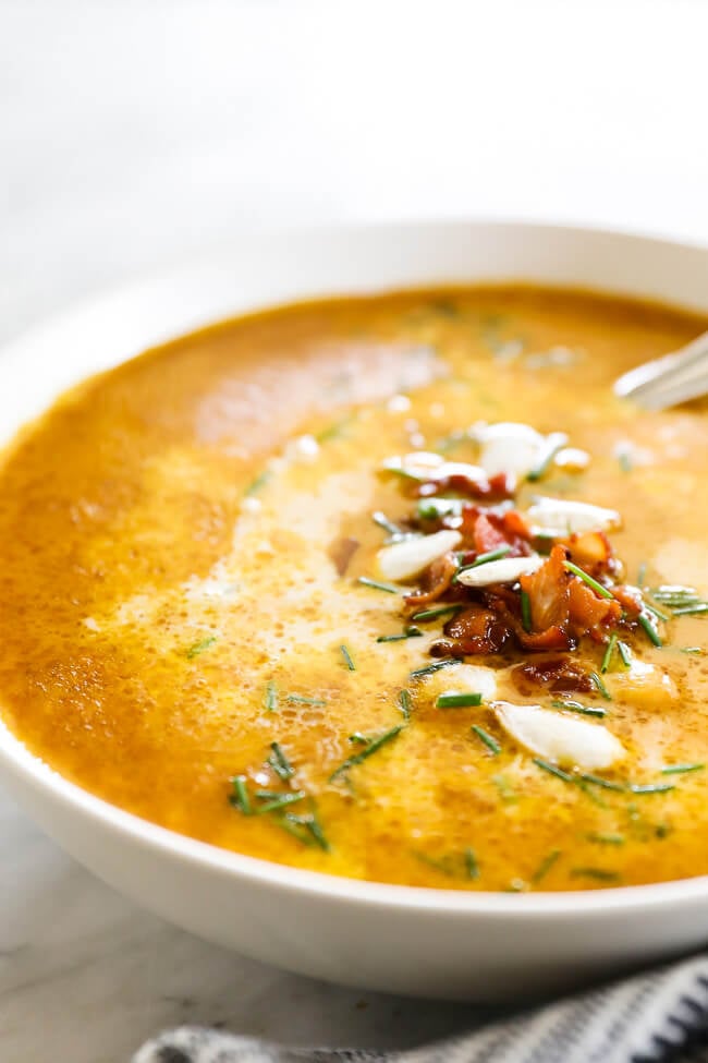 Vertical angled image of pumpkin soup in a bowl. Topped with bacon bits, pumpkin seeds and chopped chives. 