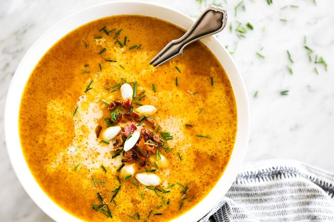 Horizontal overhead image of pumpkin soup in a bowl with a spoon. Topped with bacon bits, pumpkin seeds and chopped chives. 