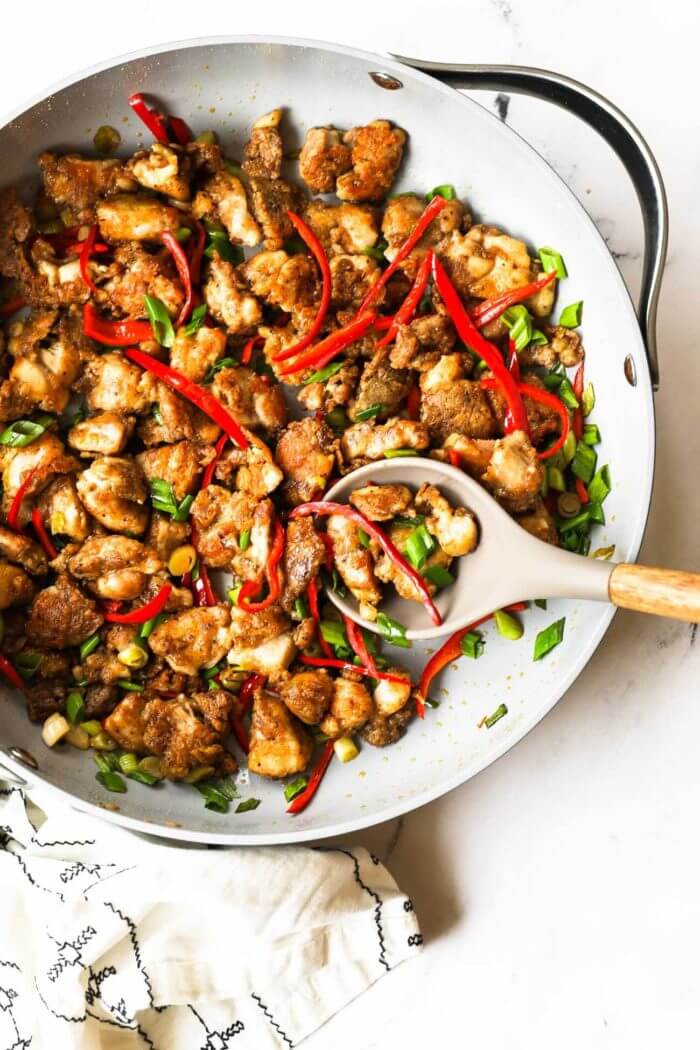 Overhead shot of easy salt and pepper chicken pieces in a skillet with red pepper strips and green onions and a spoon in the pan