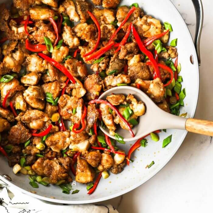 Overhead shot of easy salt and pepper chicken pieces in a skillet with red pepper strips and green onions and a spoon in the pan