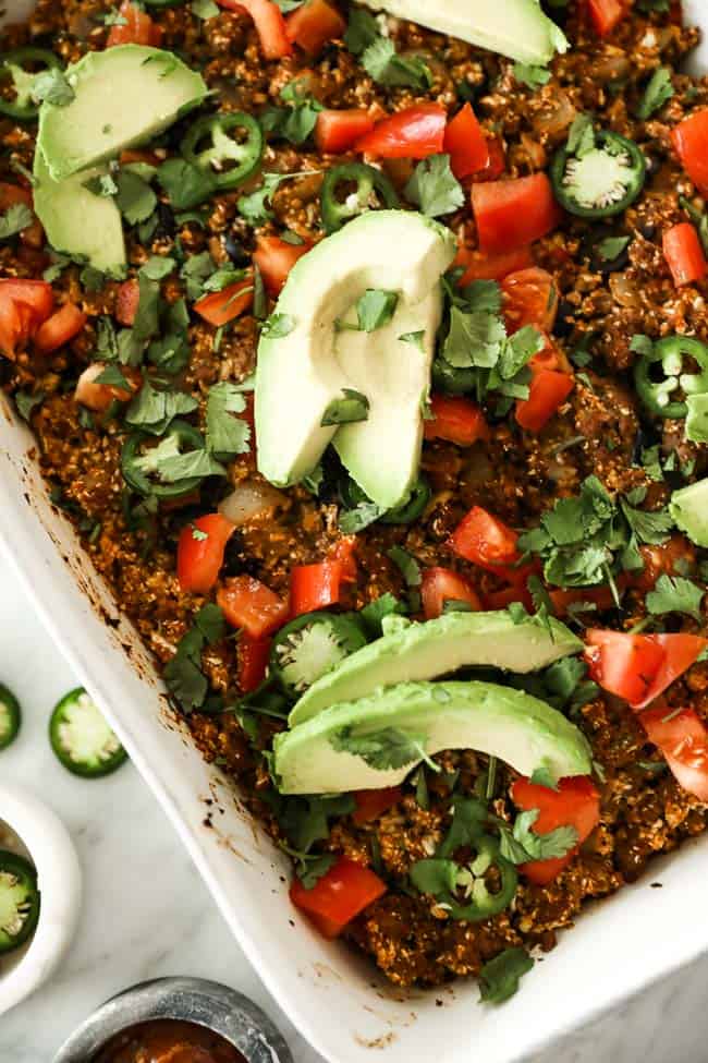 Image of easy taco casserole in white casserole dish, topped with chopped tomato, fresh cilantro, sliced jalapeño and avocado. 