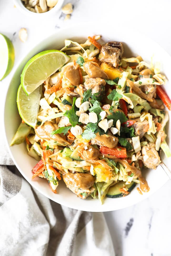 Vertical overhead image of Thai chicken salad all mixed up and served in a bowl with lime wedges, fresh cilantro and chopped cashews on top. 