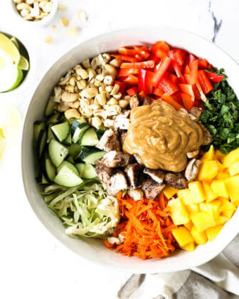 Vertical overhead image of thai chicken salad in a large serving bowl with all the ingredients separated out - cabbage, cucumber, cashews, red bell pepper, cilantro, mango, carrots, chicken and a peanut sauce.