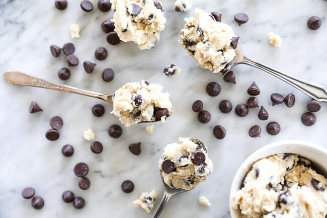Horizontal image of edible cookie dough recipe served in spoons laying out on marble with chocolate chips sprinkled around. 