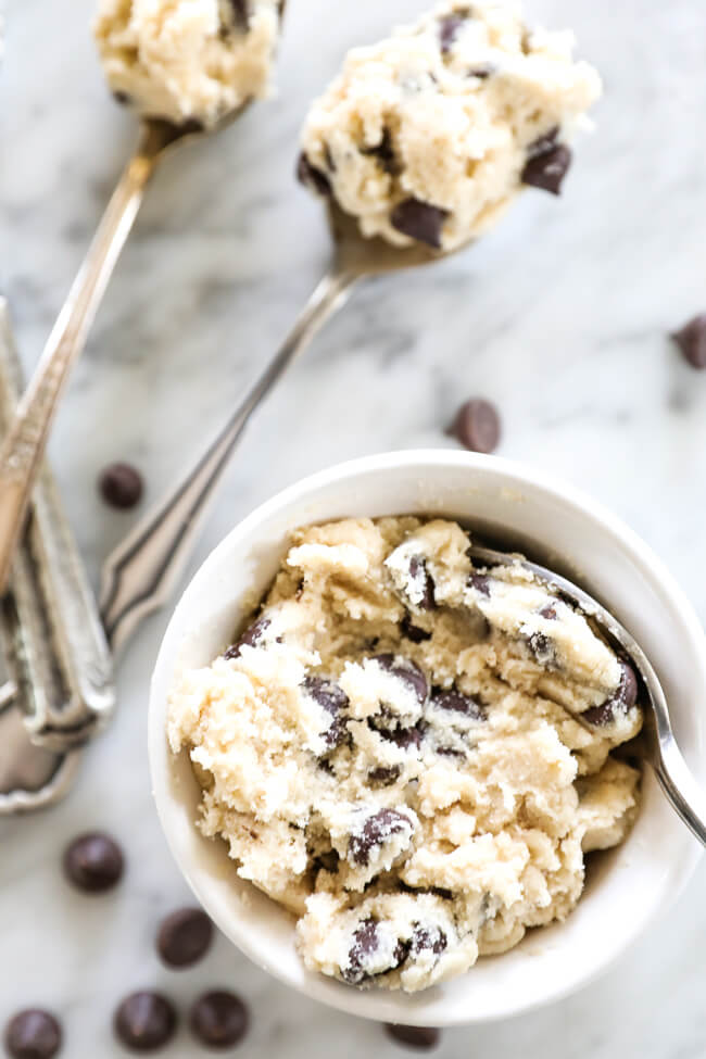 Overhead vertical image of edible cookie dough in a ramekin with a spoon dipped in. Chocolate chips and a couple spoonfuls off to the side. 