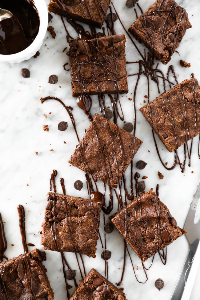 Vertical image of squares of egg-free brownies laid on out on marble with chocolate sauce and extra chocolate chips drizzled and sprinkled all around. 