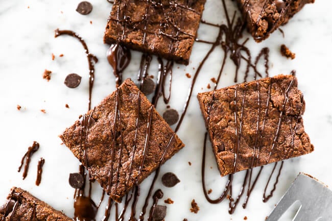 Horizontal image of egg-free brownies cut into squares and laid on marble with chocolate sauce drizzled on tp and chocolate chips spread around. 