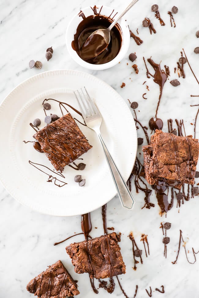 Vertical image of egg-free brownies. Some on marble and one square on a plate with a fork. Chocolate sauce drizzled all around and extra sauce on the side in a small ramekin with a spoon. 
