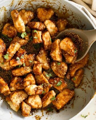 Close up overhead image of cooked crispy honey sesame chicken pieces in a pan with spoon and topped with sesame seeds