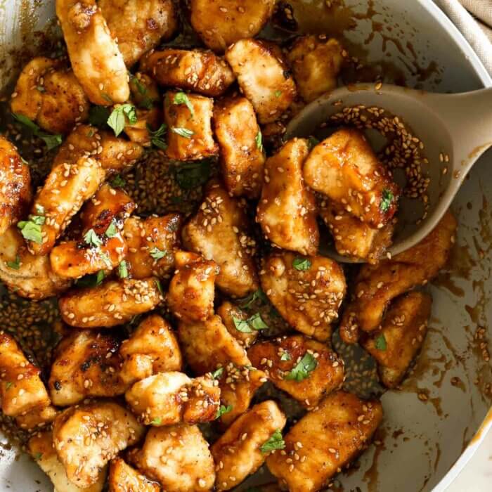 Close up overhead image of cooked crispy honey sesame chicken pieces in a pan with spoon and topped with sesame seeds