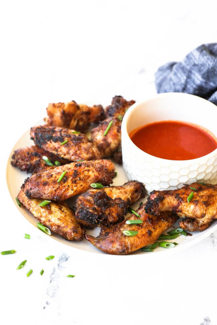 Angled image of crispy chicken wings piled on a plate with buffalo sauce in a ramekin on the side.