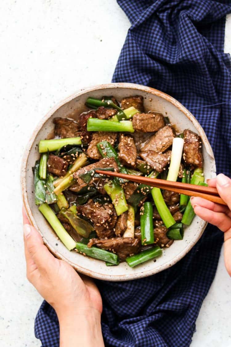 Mongolian beef on a plate with green onions, sesame seeds and hands with chopsticks.