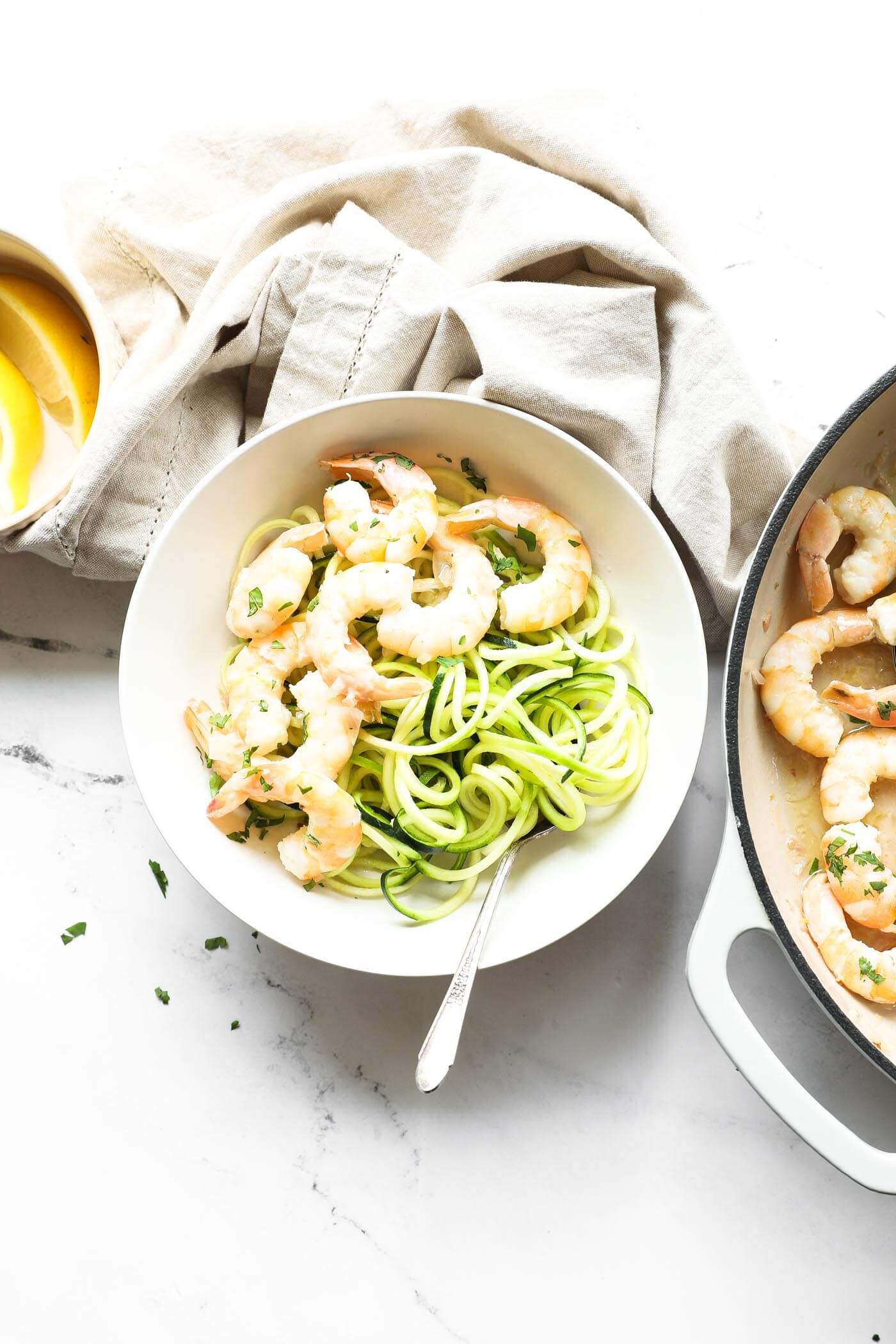 Overhead image of a garlic butter shrimp served over zoodles in a bowl with a fork. Shrimp is sprinkled with chopped cilantro on top. Extra shrimp and lemon wedges on the side.