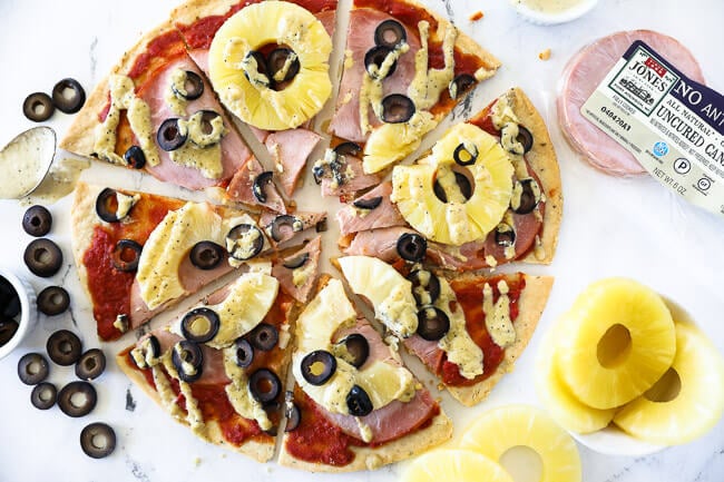 Horizontal image of hawaiian pizza cut into slices with olives, pineapple, canadian bacon, marinara sauce and a creamy dairy free sauce drizzled on top. 
