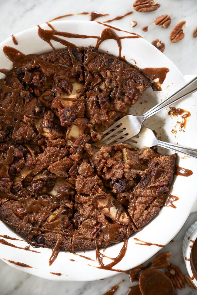 Close up of gluten-free apple cake in a pie dish with piece removed and two forks dug in. Caramel sauce drizzled all over top. 