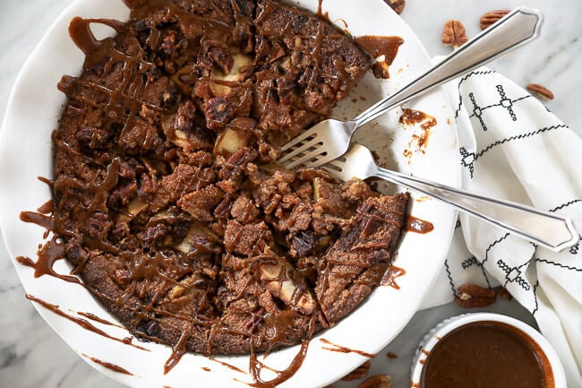 Gluten-free apple cake in a pie dish with a piece taken out and two forks dug. Caramel sauce drizzled all over the top.  