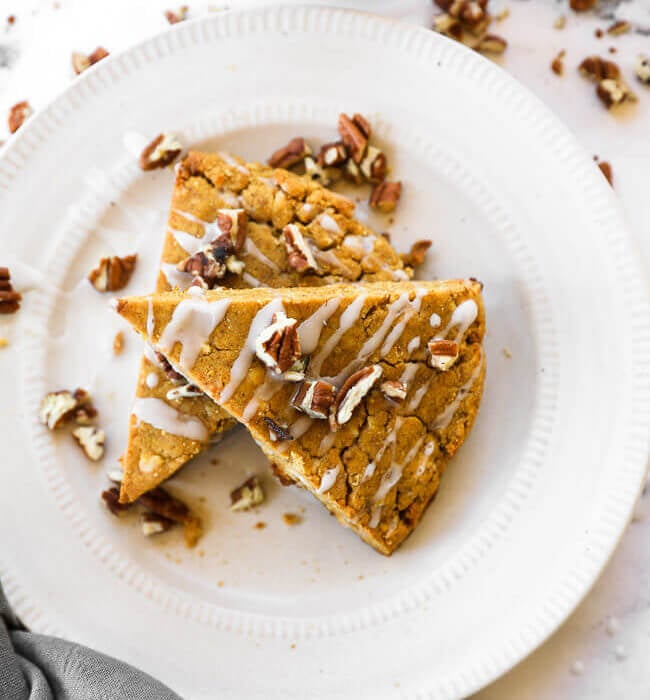 Vertical image of two gluten free and vegan pumpkin scones stacked on a plate with icing and chopped pecans on top.