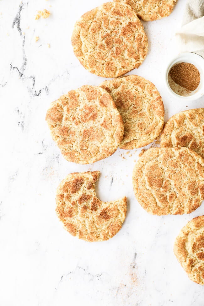 Vertical overhead image of homemade snickerdoodles laid out on marble. One cookie has a bite taken out of it. 