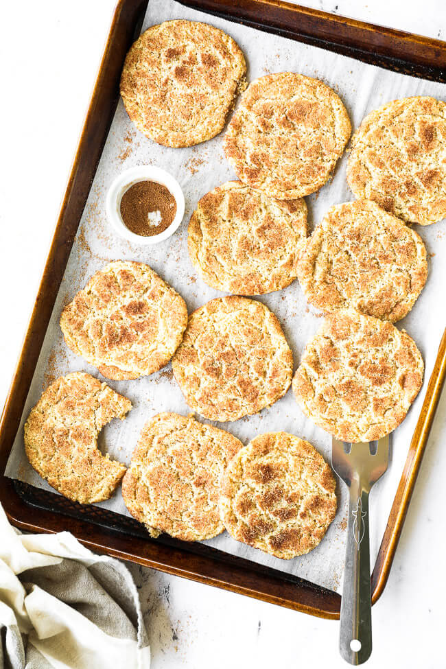 Overhead vertical image of homemade snickerdoodles on a sheet pan with extra cinnamon and sugar and a spatula under one cookie. 