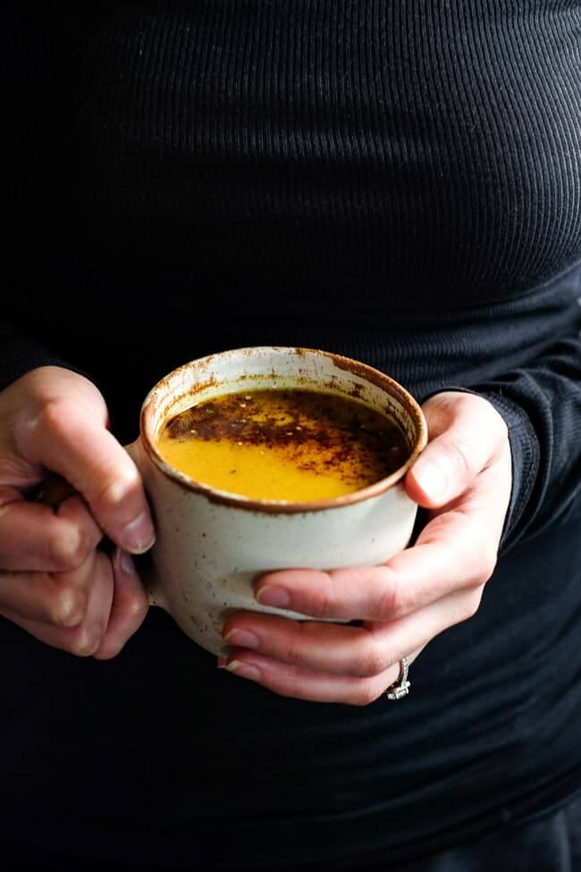 Holding a mug of golden milk latte. It's topped with cinnamon and hemp seeds. 