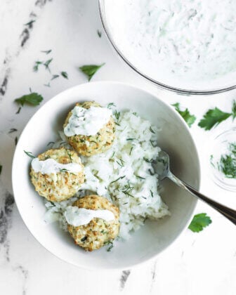 A white bowl with three meatballs on a bed of rice, topped with white sauce and herbs, with a spoon and a bowl of extra sauce beside it on a marble surface.