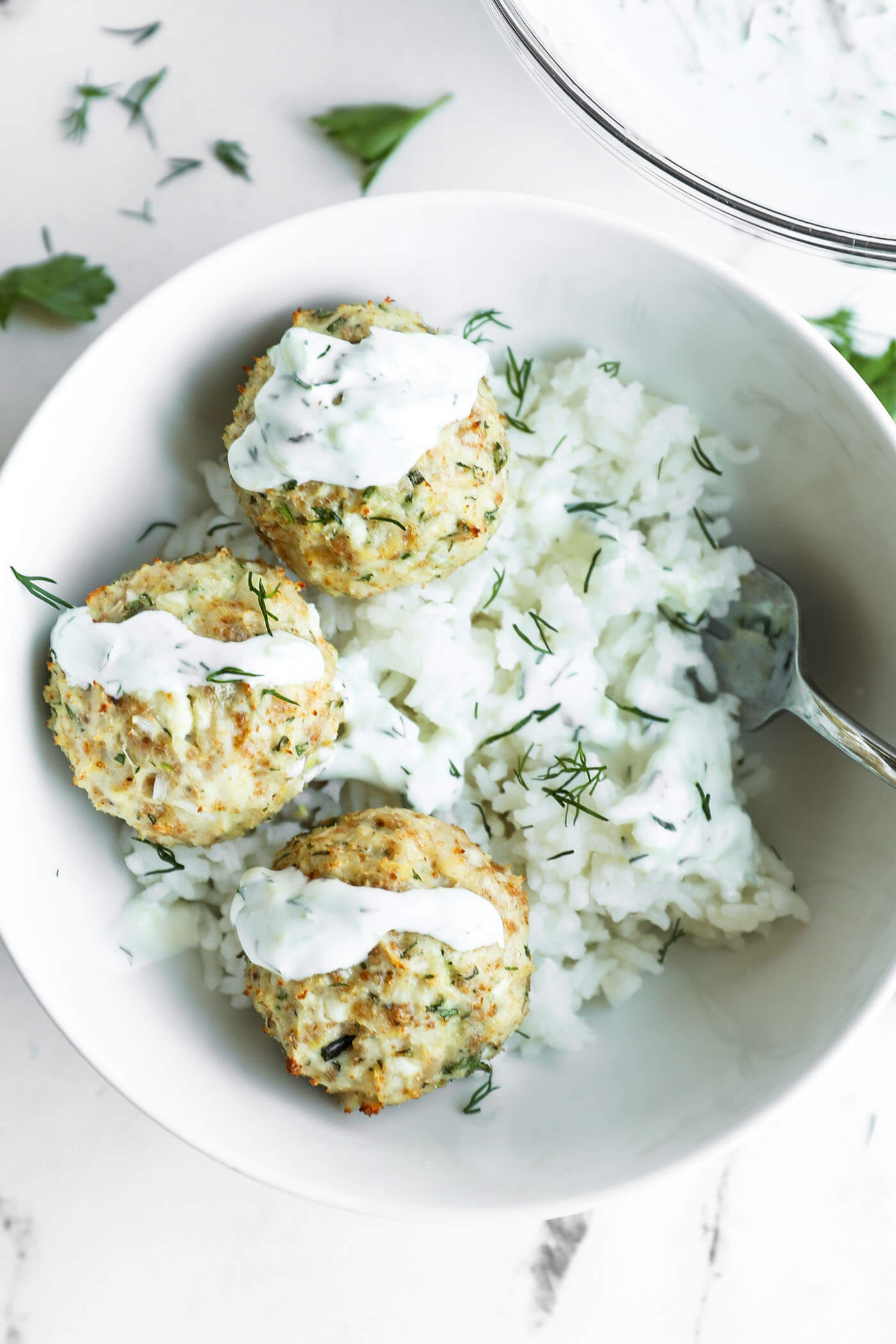 Three Greek chicken meatballs with white sauce served on a bed of rice, garnished with fresh dill, in a white bowl with a fork.