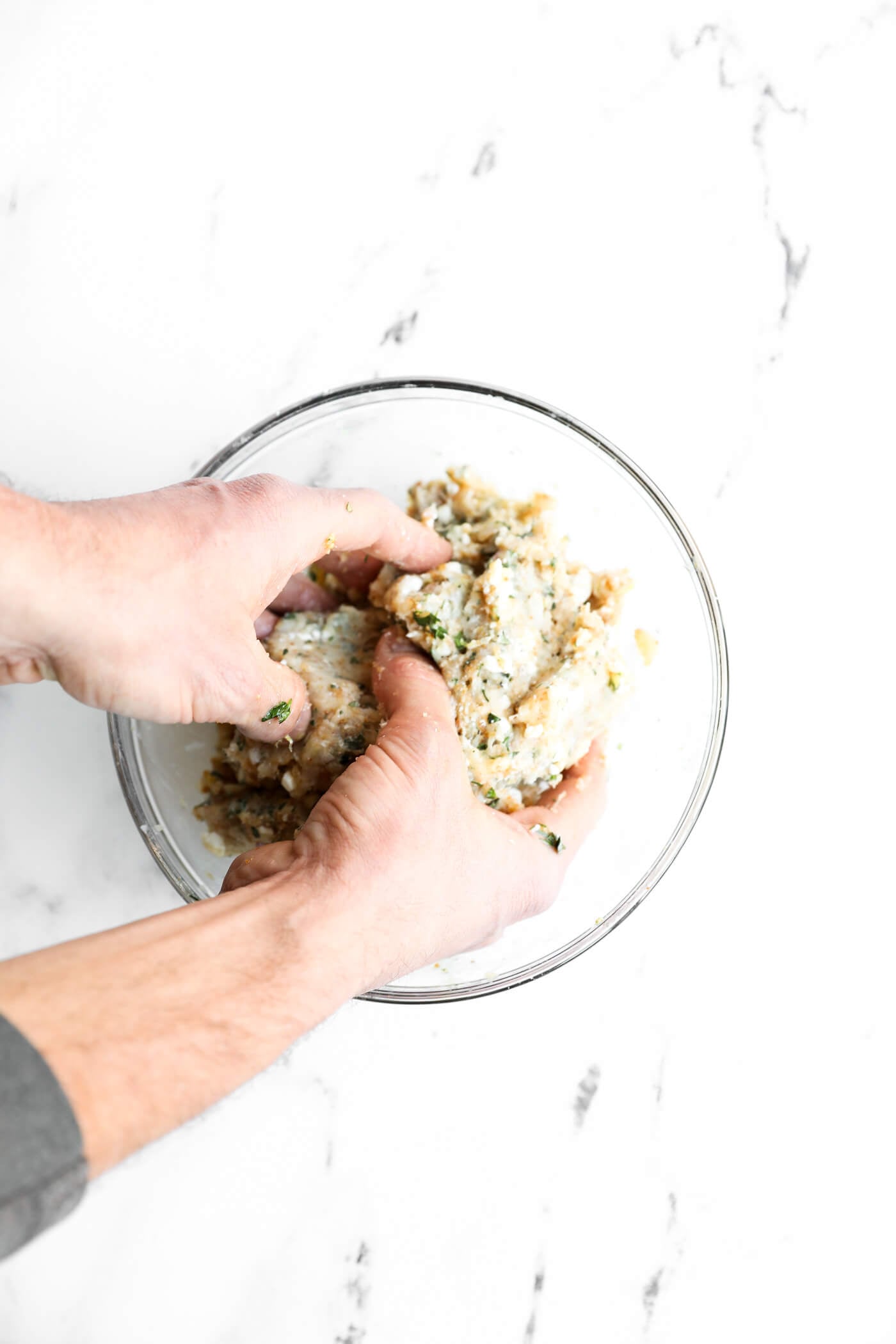 Two hands mixing a mixture of herbs, breadcrumbs, and ground meat in a clear glass bowl on a white marble surface.