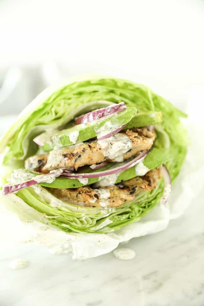 Two Greek turkey burgers stacked between lettuce buns. Topped with avocado and red onion and drizzled with tzatziki sauce. 