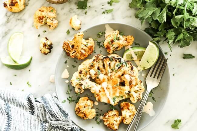 Grilled cauliflower on a plate topped with creamy sauce, chopped cilantro and lime wedges with a fork. 