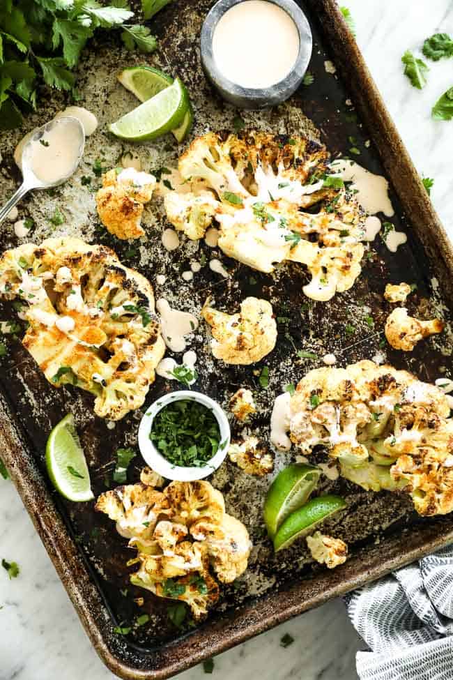 Cauliflower steaks spread out on a baking sheet with cream sauce drizzled on top and topped with cilantro. Cilantro, lime wedges, sauce and a spoon on pan. 