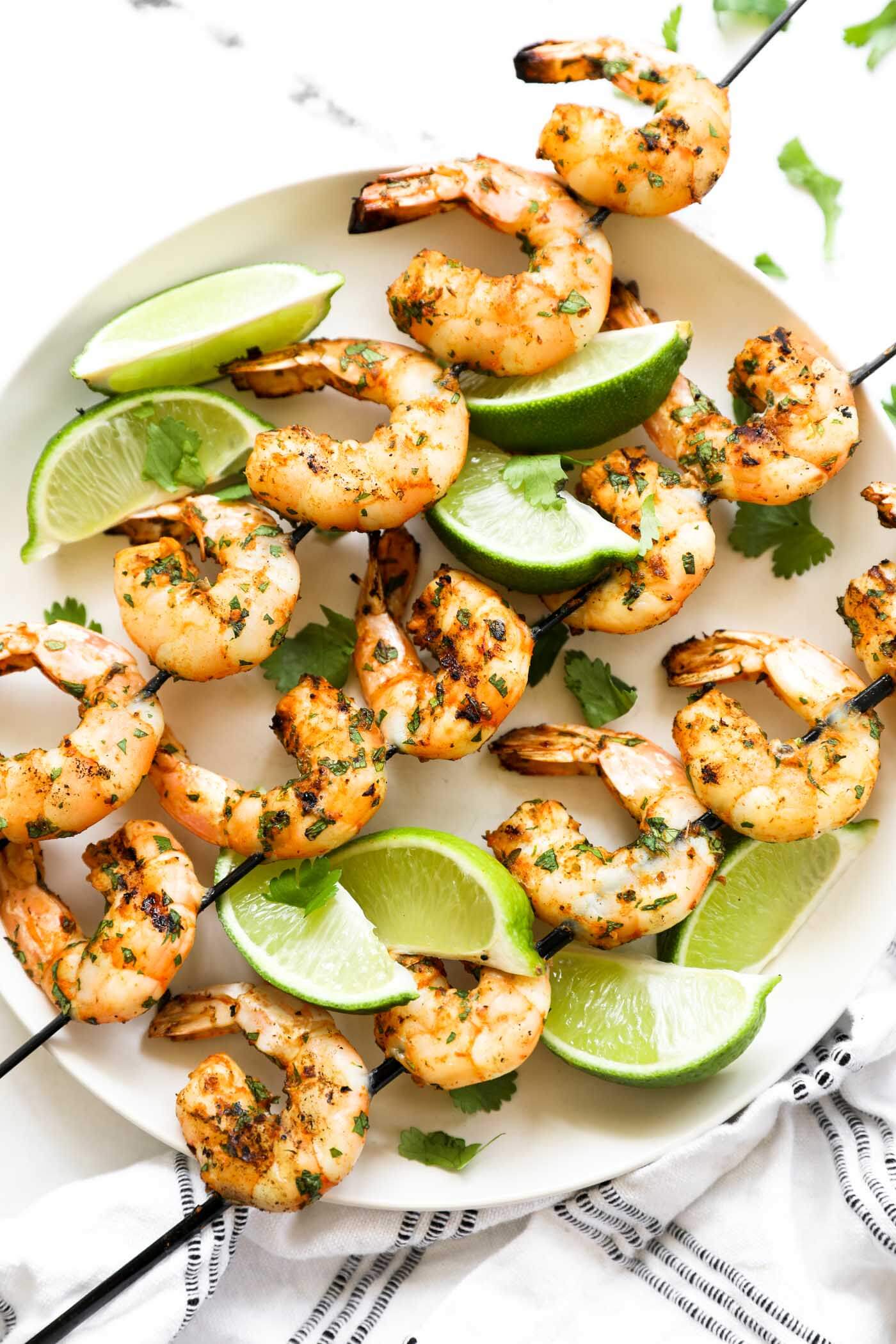 Grilled shrimp kabobs laid out on a plate. 3 skewers with lime wedges and fresh cilantro garnish.