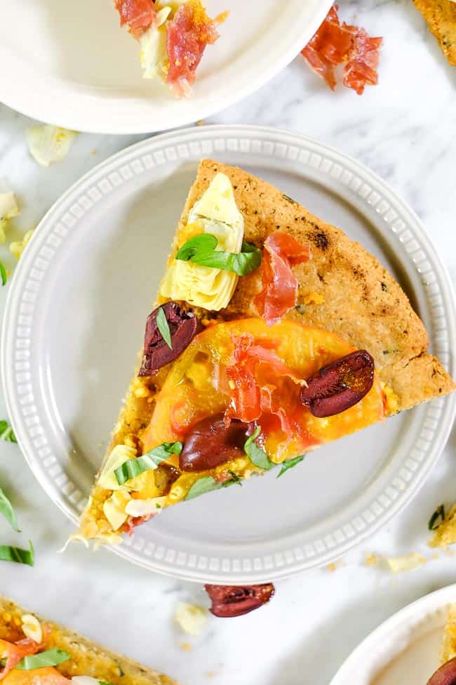 Close up of grilled paleo pizza with heirloom tomatoes, artichoke hearts, kalamata olives, prosciutto and fresh basil on a plate. 