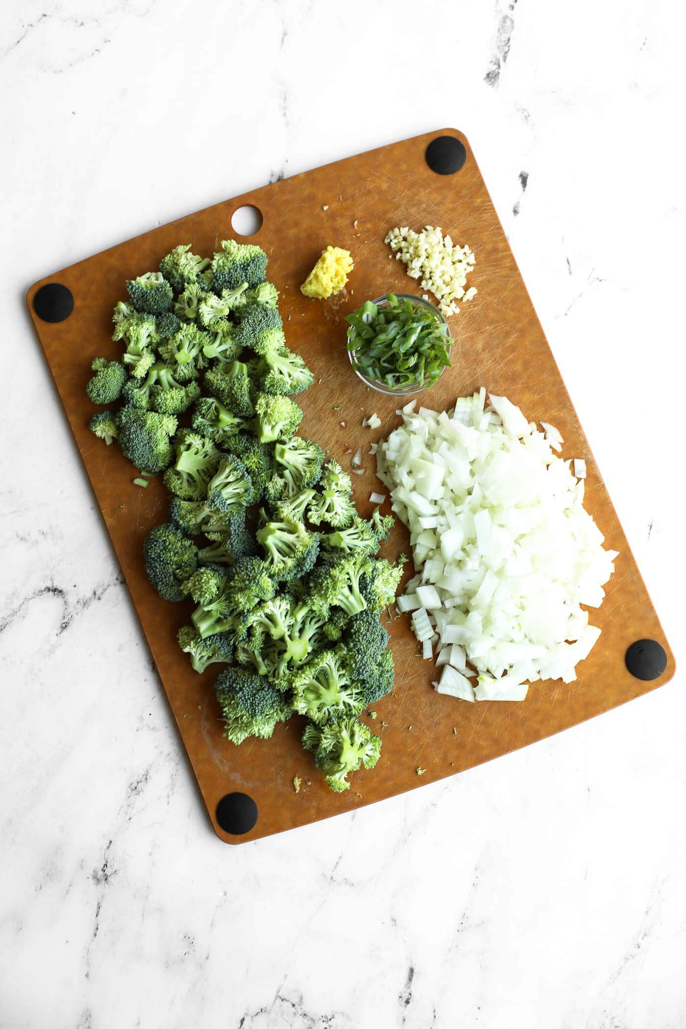 Chopped broccoli, diced onion, sliced green onions, minced garlic, and grated ginger on a brown cutting board.