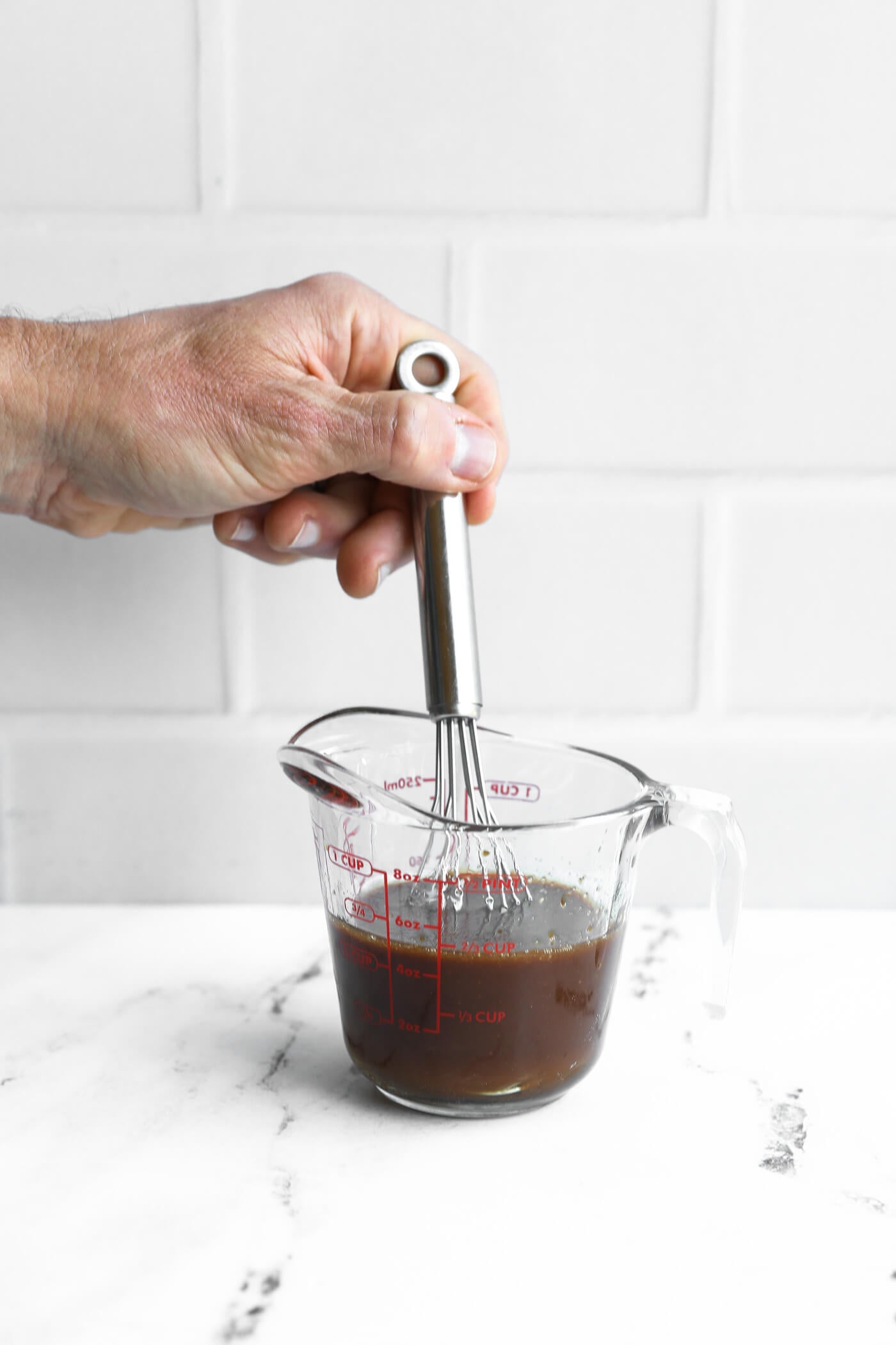 A hand holds a whisk in a glass measuring cup filled with dark liquid on a white countertop with a white tile background.