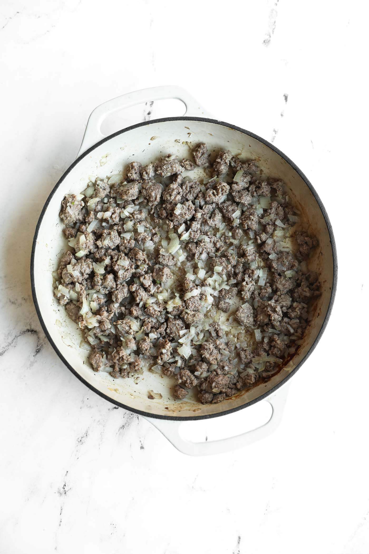A white pot on a marble surface contains cooked ground beef mixed with chopped onions.