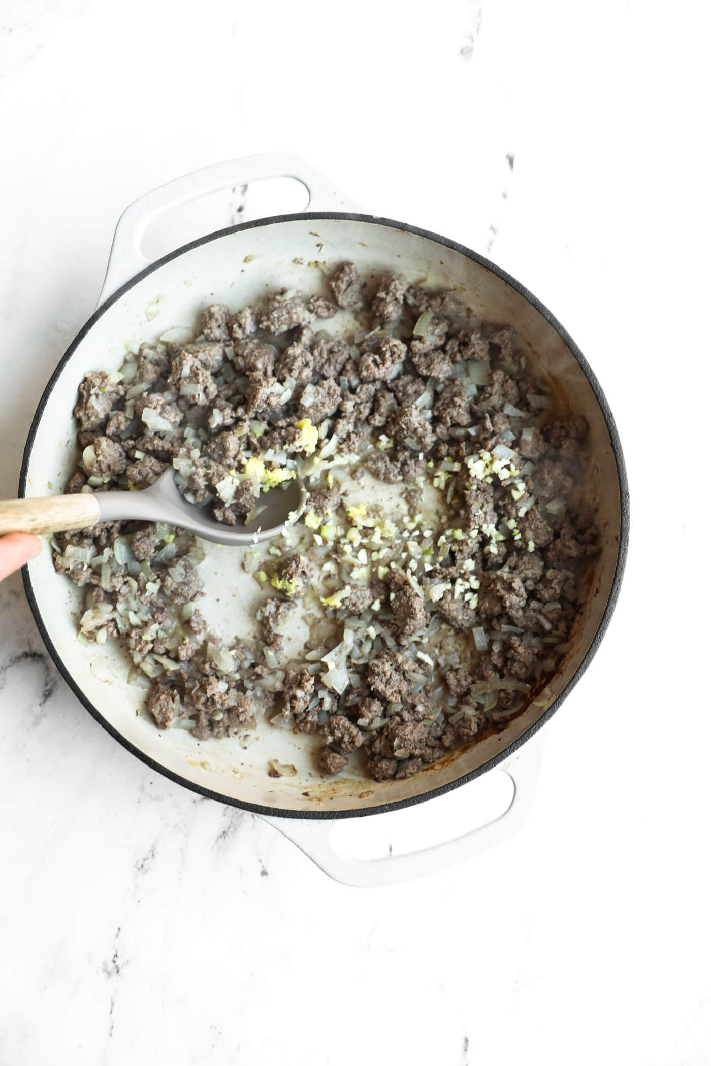Ground meat, chopped onions, and minced garlic are being sautéed in a white pot on a white surface, with a hand stirring using a wooden spoon.